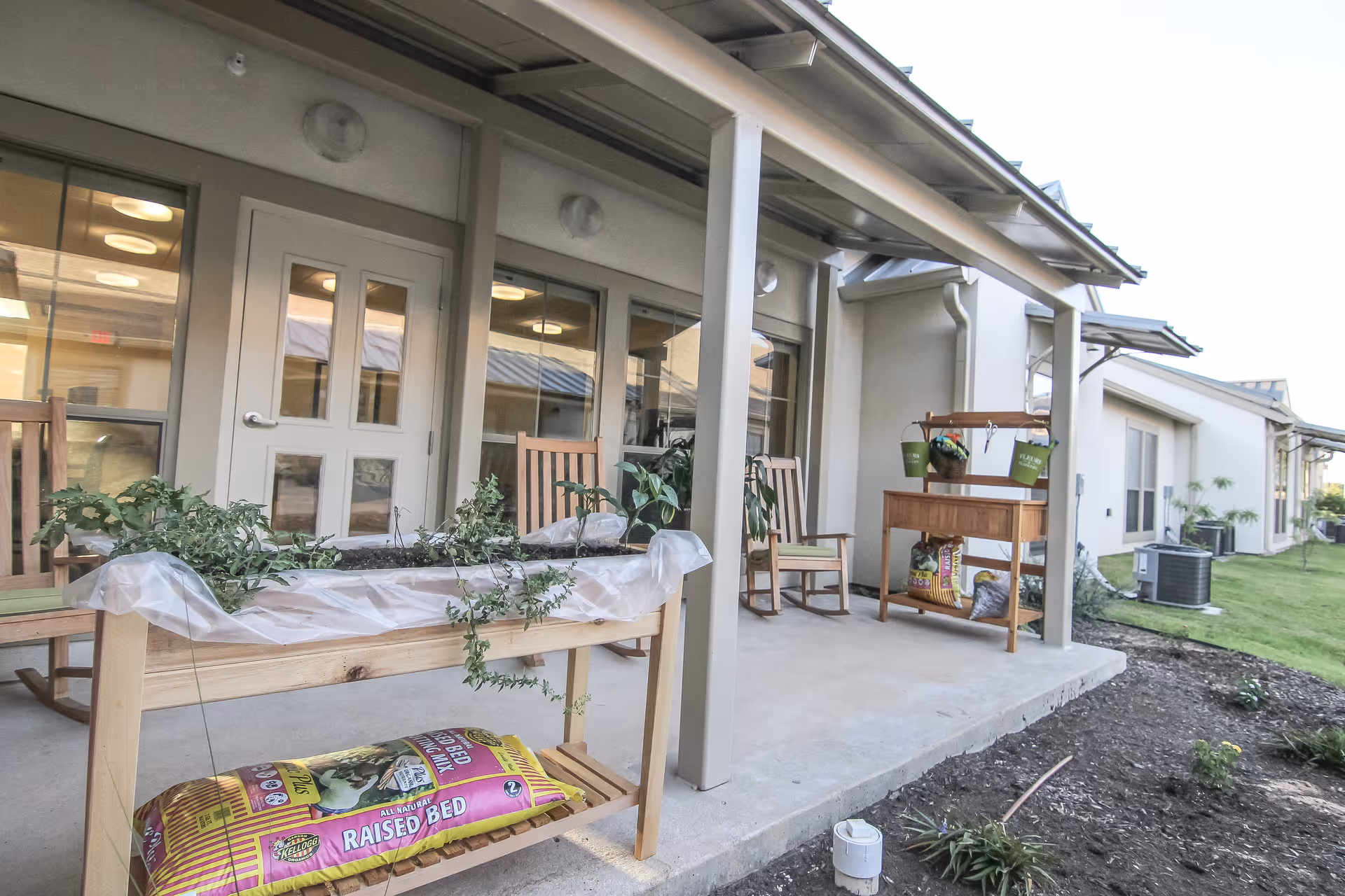 Outdoor covered patio area with wooden rocking chairs, raised garden beds with plants, and gardening supplies. The patio is adjacent to a building with large windows and doors, and there is a grassy area and garden beds beside the patio.