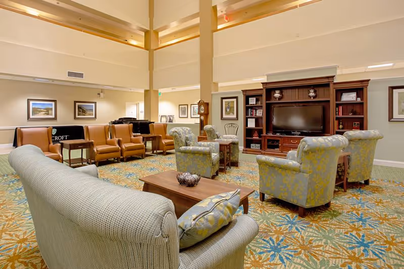 Spacious senior living common room with sofas and armchairs arranged around a large TV cabinet.