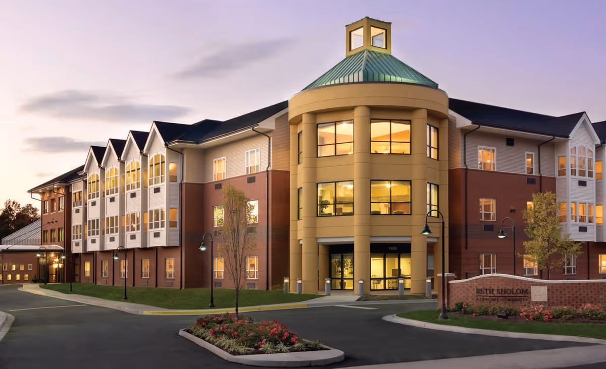 Exterior view of a multi-story senior living facility building at dusk with warm lights glowing from the windows. The building features a prominent rounded entrance with large windows and a green roof. There are street lamps, a small landscaped area with flowers and trees, and a sign that reads 'Beth Sholom A Lifecare Community' near the entrance.