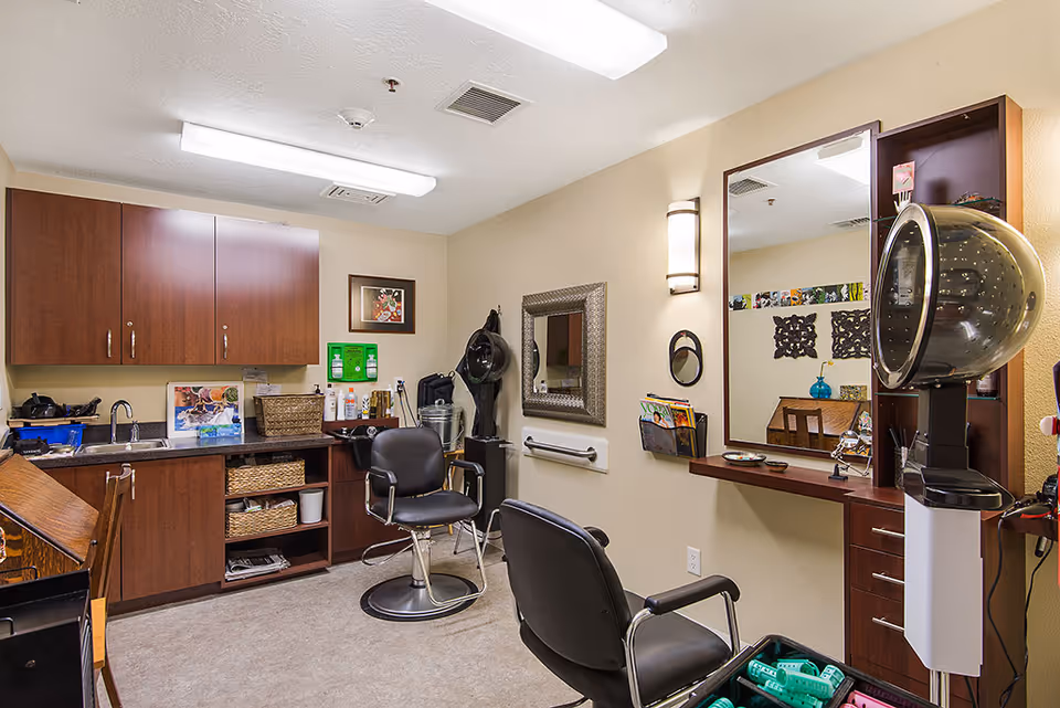Interior view of a small salon room in an assisted living facility featuring two black salon chairs, a hair dryer hood, a large mirror, wooden cabinets, and various hair care supplies on the counter and shelves.