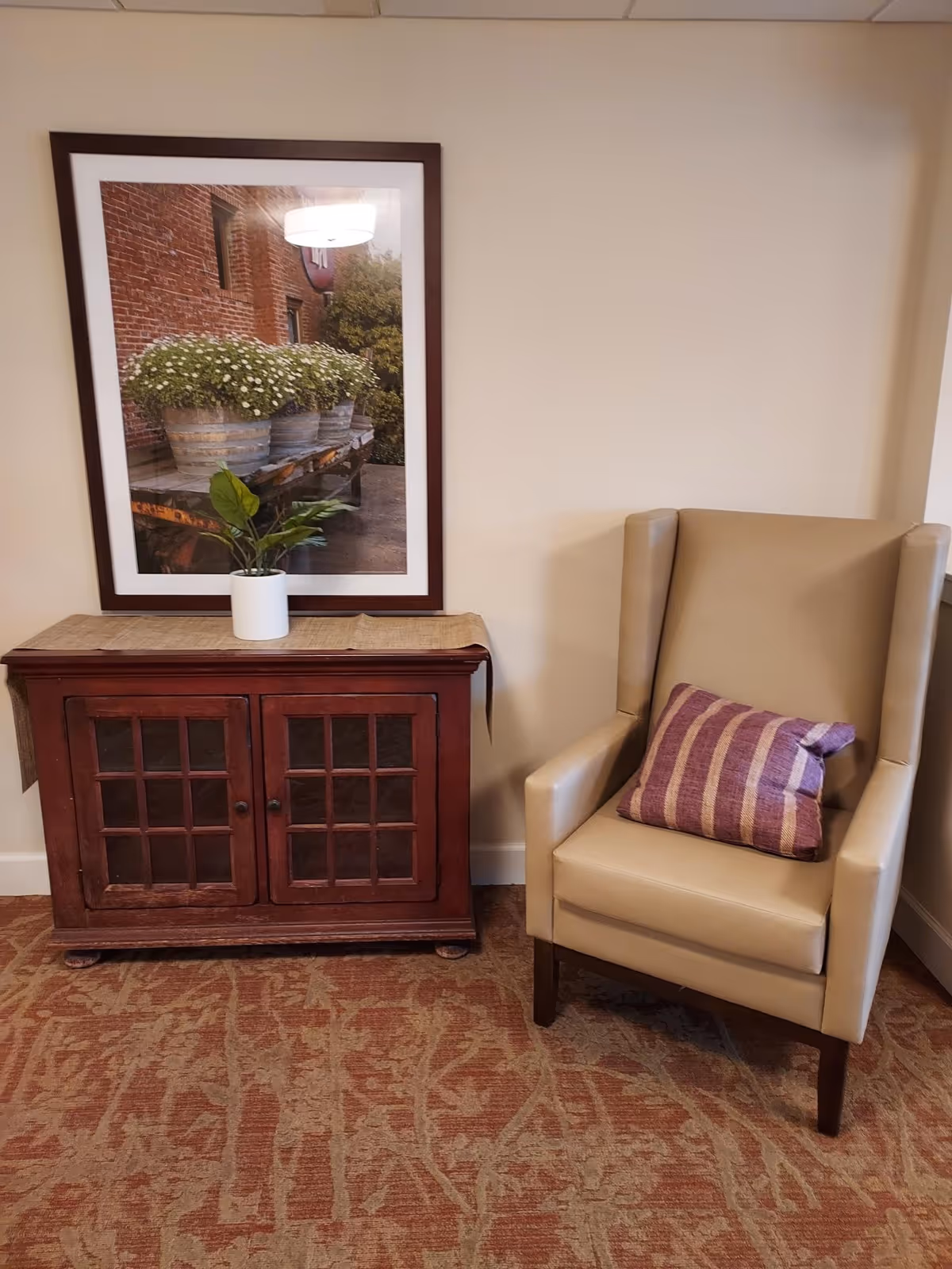 A beige leather armchair with a purple striped pillow is positioned next to a wooden cabinet with glass-paneled doors. On top of the cabinet is a small potted plant and a framed picture of flower pots on a wooden table hanging on the wall above. The floor is covered with a patterned carpet.