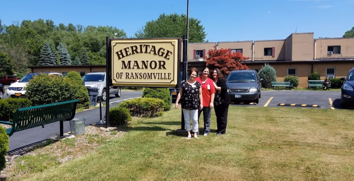 Three women standing on a grassy area in front of a sign that reads 'Heritage Manor of Ransomville' with a parking lot and a building in the background under a clear blue sky.