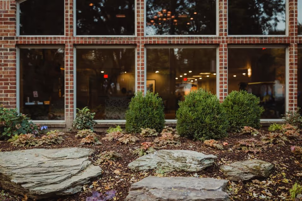 View of a brick building exterior with large windows reflecting trees and showing a warmly lit interior. In front of the building, there is a landscaped garden area with rocks, small bushes, and plants.