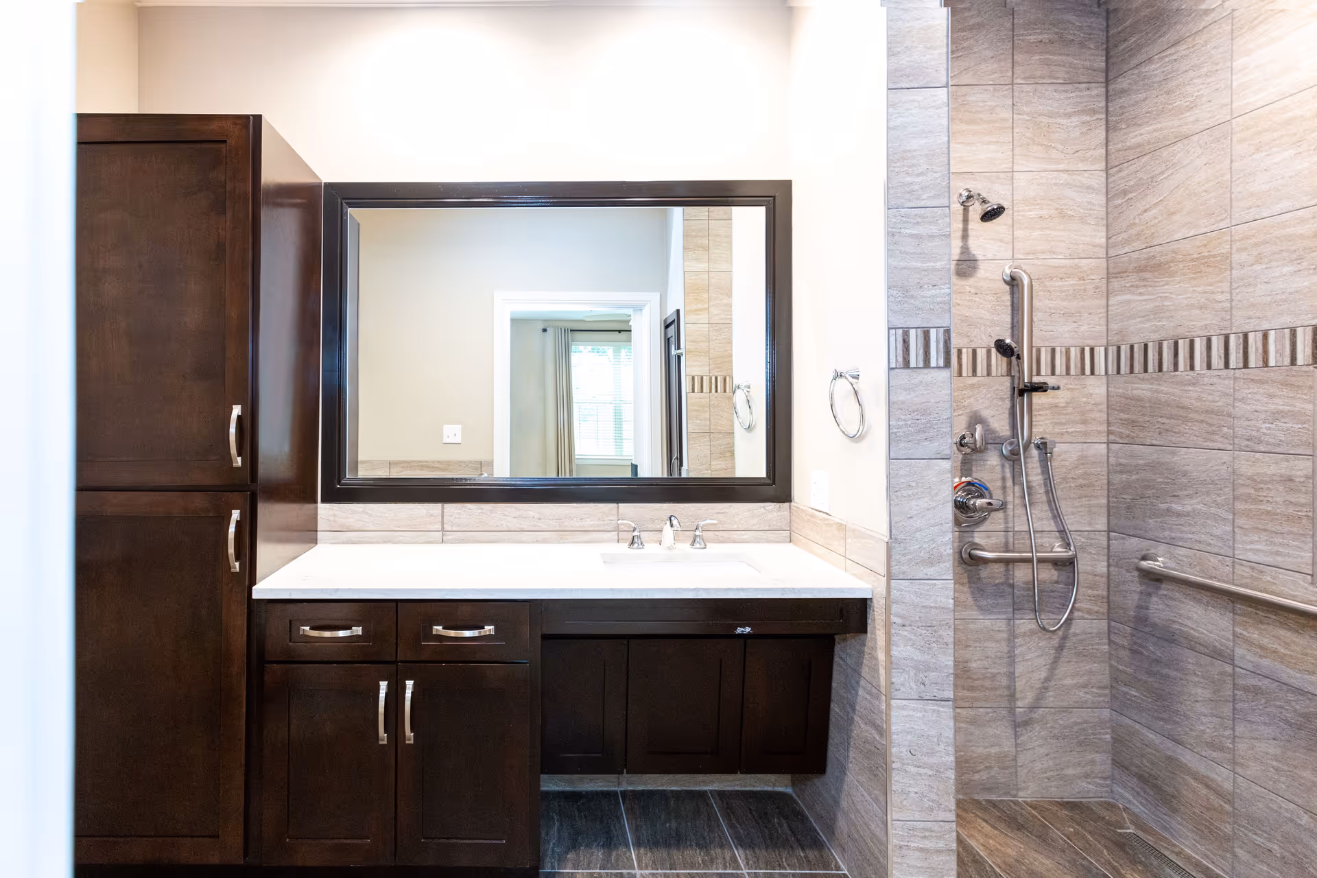 Bright bathroom featuring a dark wood vanity with a white countertop and large mirror beside a tiled walk-in shower with grab bars and a handheld showerhead.