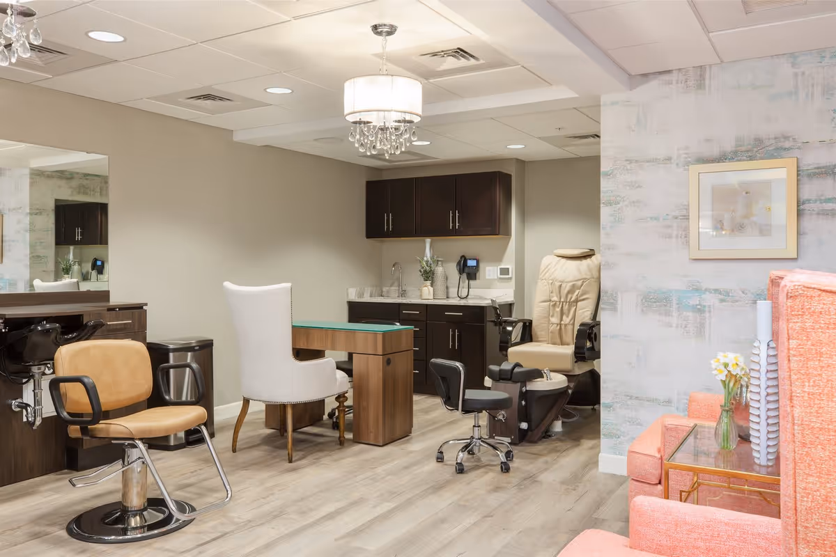 Interior view of a salon area in a senior living facility featuring salon chairs, a manicure table with a white upholstered chair, a pedicure chair, dark wood cabinetry with a sink, and a small seating area with coral-colored chairs and a glass side table with flowers.