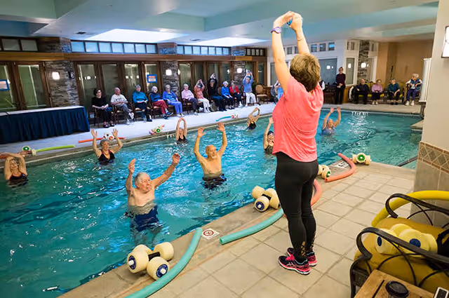 A group of elderly people participating in a water exercise class in an indoor swimming pool, led by an instructor standing poolside with arms raised. Other seniors are seated along the wall watching the activity.