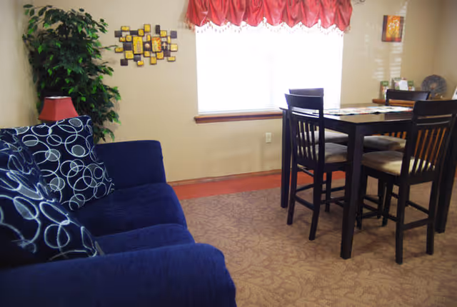 A cozy interior room featuring a dark blue couch with patterned cushions on the left and a wooden dining table with four chairs on the right. A window with a red valance lets in natural light, and the walls are decorated with a plant and framed artwork.