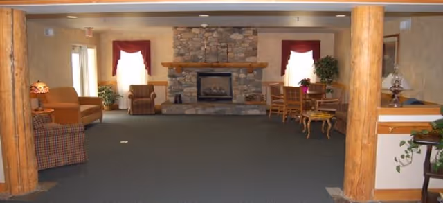 Spacious common room with a stone fireplace centered between two curtained windows, seating, tables, and exposed wooden columns.