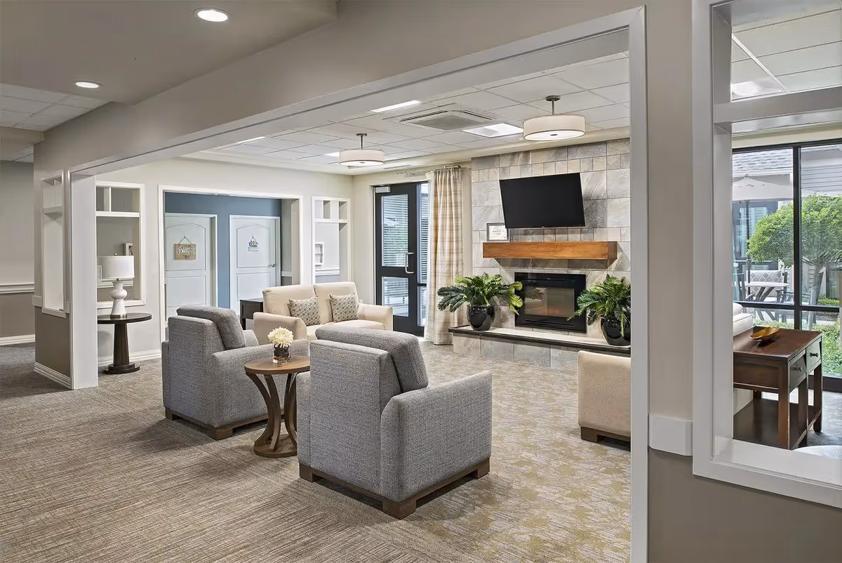 A cozy living room area in a senior living facility featuring two gray armchairs and a beige loveseat arranged around a small wooden table with a flower vase. The room has a modern fireplace with a mounted flat-screen TV above it, flanked by two potted plants. Large windows and glass doors let in natural light and offer a view of an outdoor patio with seating and greenery. The walls are painted in neutral tones, and the carpet has a subtle pattern. There is a side table with a lamp near the entrance to another room with two white doors.