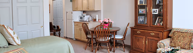 A cozy living space featuring a bed with pillows on the left, a small round wooden dining table with four chairs in the center, a kitchenette with cabinets, a microwave, and a coffee maker in the background, and a wooden china cabinet with framed photos and decorative items on the right. There is also a patterned armchair partially visible on the right side.