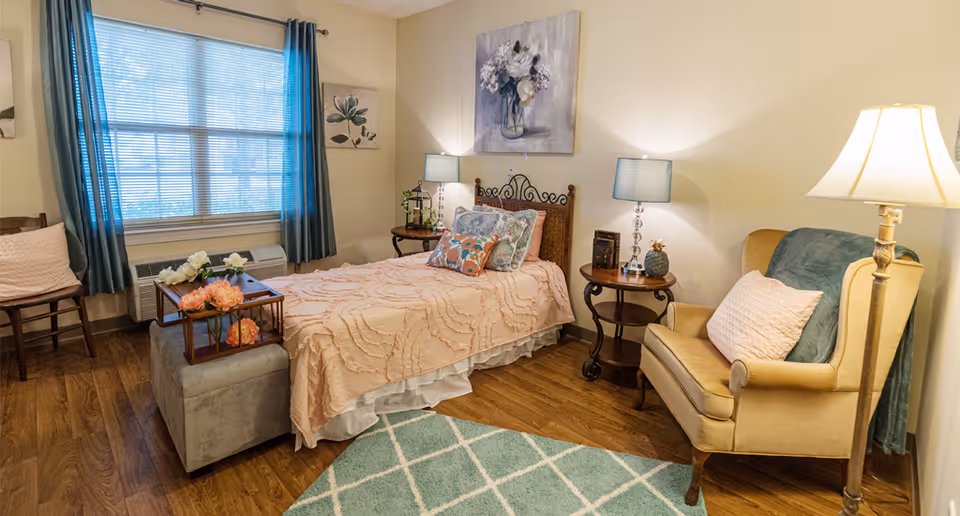 Well-lit furnished bedroom with a single bed, armchair, side tables, lamps, window and decorative accents.