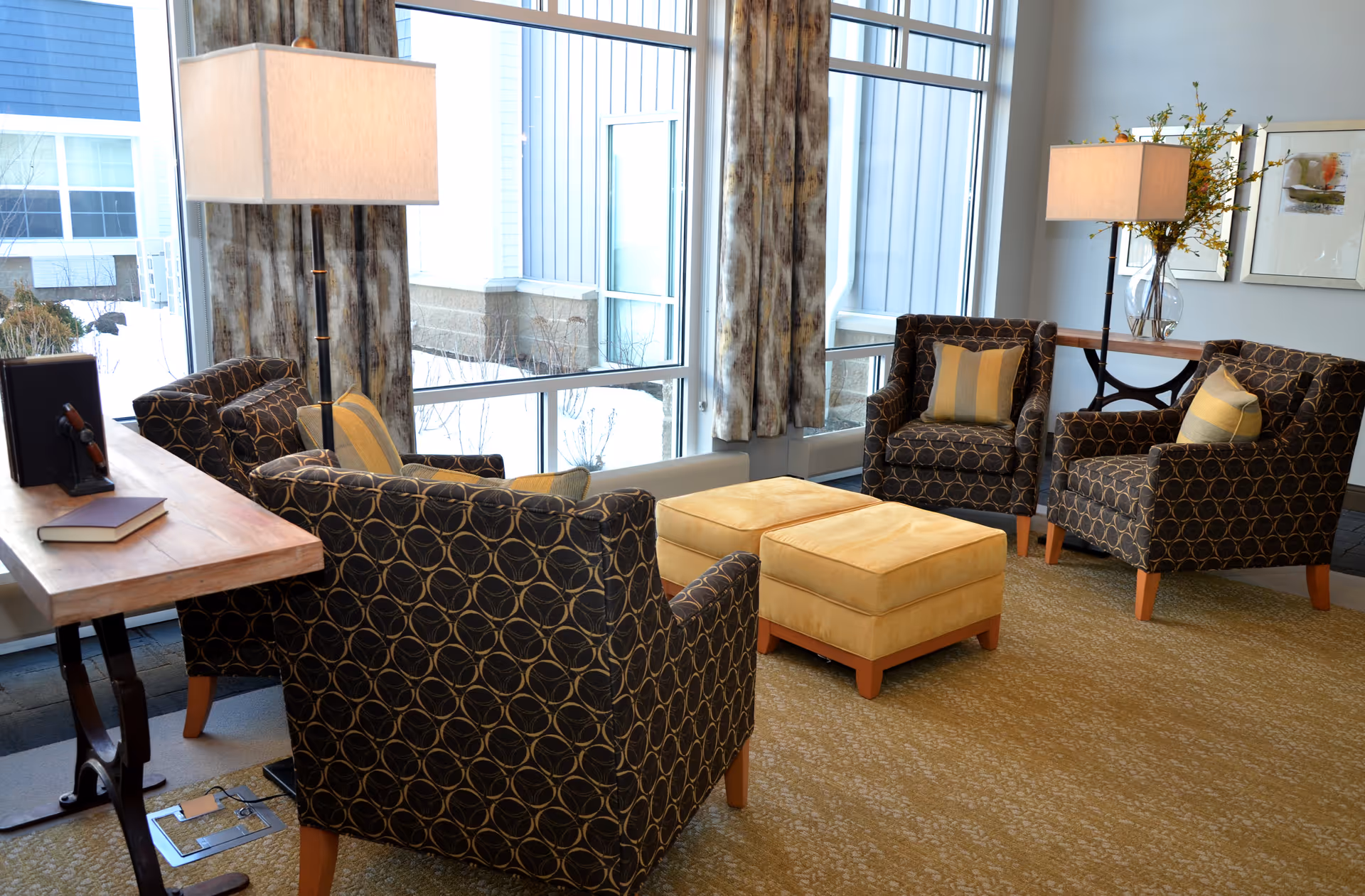 A cozy sitting area with four patterned armchairs arranged around two yellow ottomans. The space is well-lit by natural light coming through large windows with curtains. There are two floor lamps with rectangular shades, a wooden table with books and a decorative item, and a console table with a vase of flowers and framed artwork on the wall.