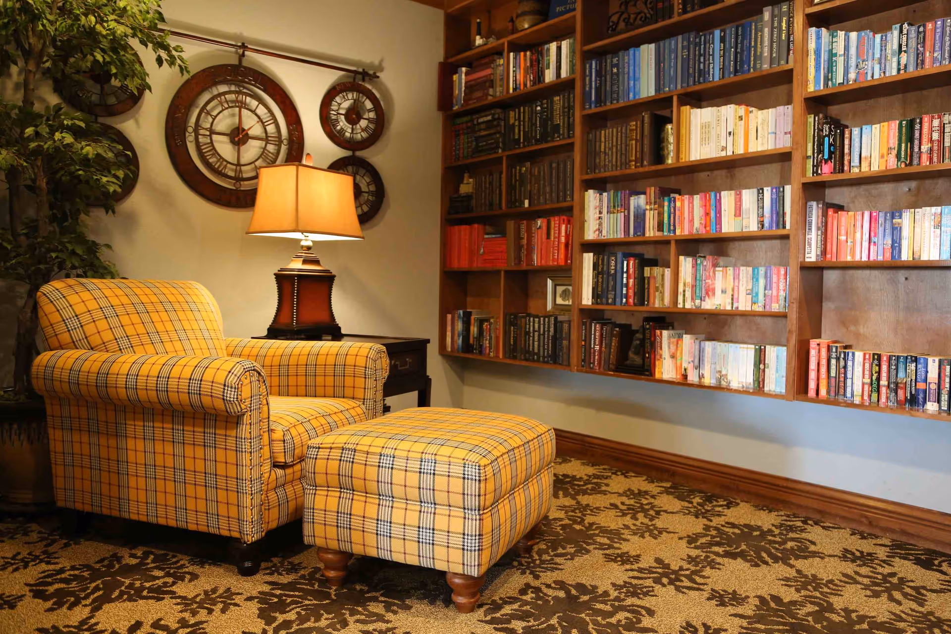 Cozy reading nook with a yellow plaid armchair and matching ottoman, a side table with a lamp, a large wall clock, and a wooden bookshelf filled with books.