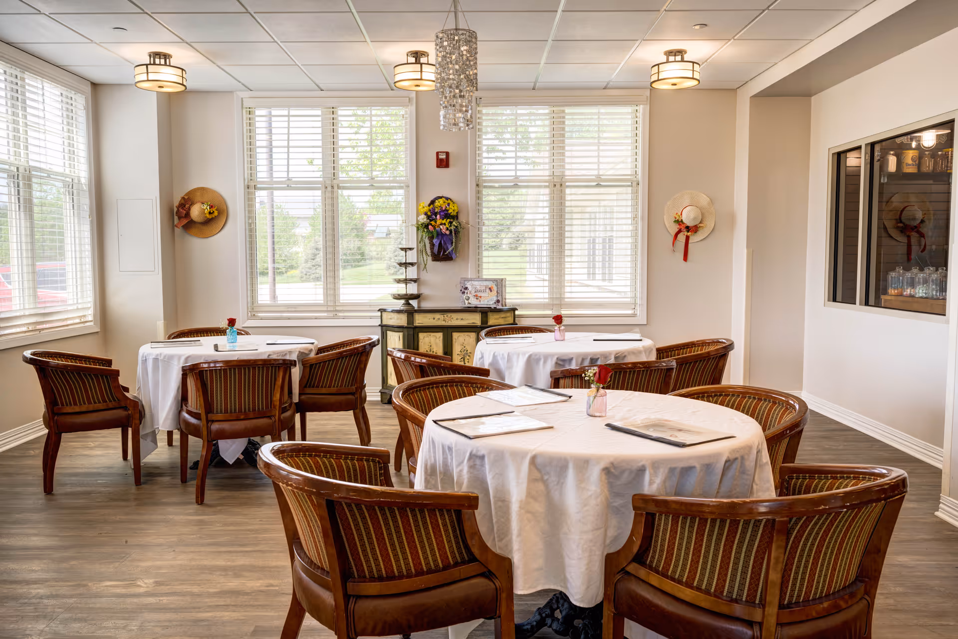 Bright dining room with round tables draped in white tablecloths and wooden chairs arranged near large windows.