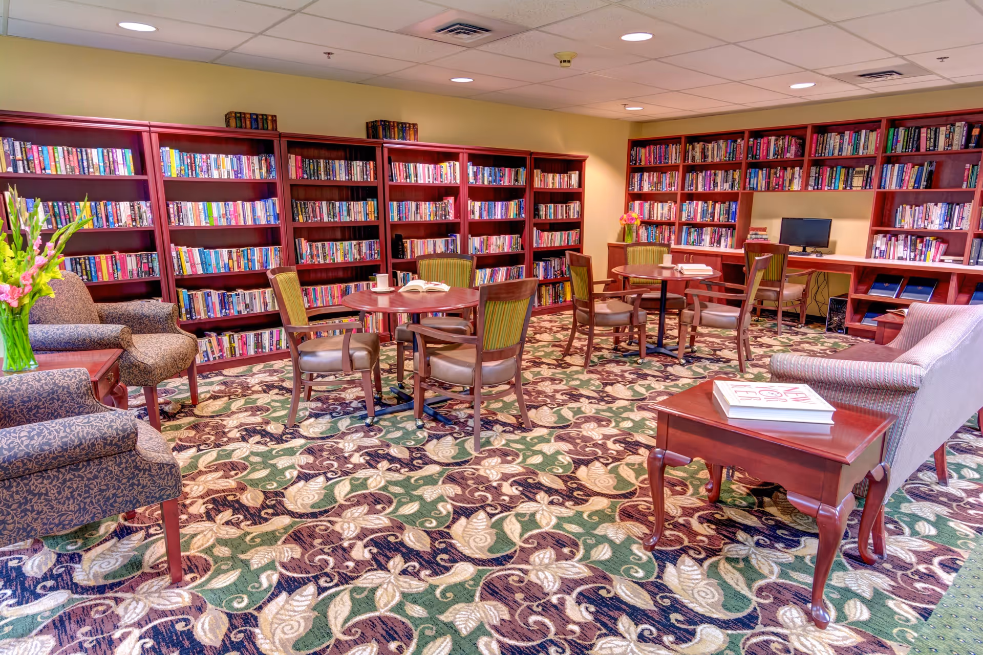 A cozy library room in Rosewood Park featuring multiple wooden bookshelves filled with books along the walls. There are round tables with chairs arranged for reading or socializing, a computer workstation, and comfortable upholstered armchairs and a sofa. The room has a patterned carpet with green, beige, and brown tones and soft yellow walls.