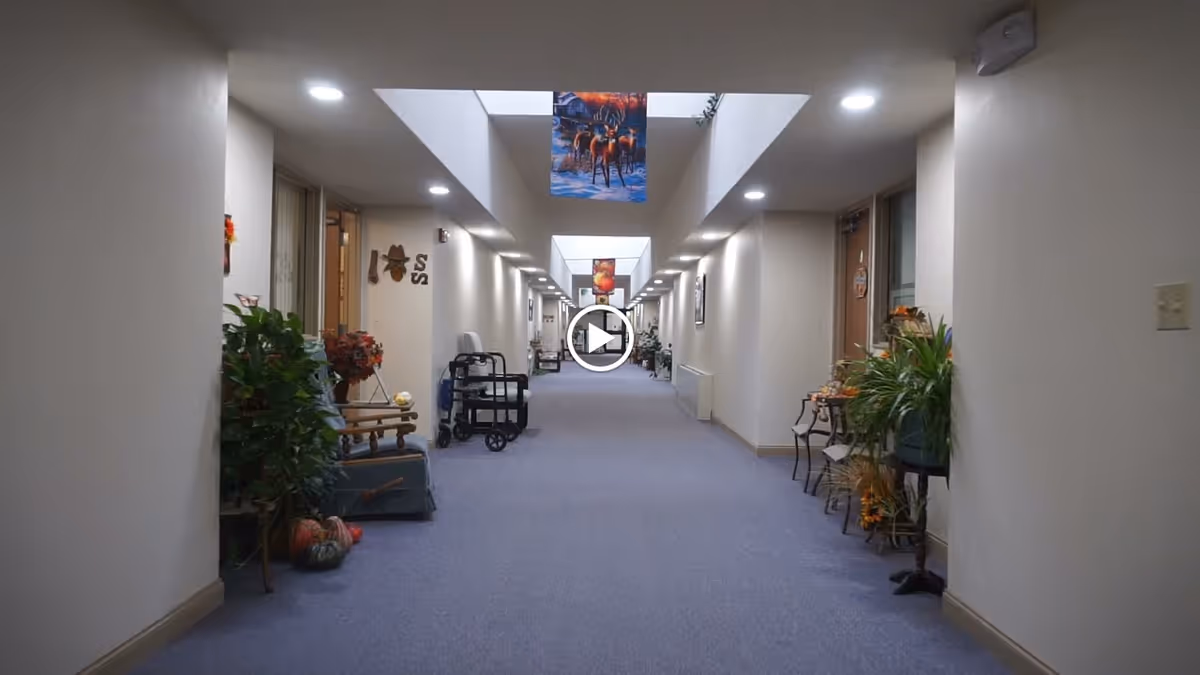 A long, well-lit hallway in an assisted living facility with light-colored walls and carpeted floor. The hallway is decorated with plants, chairs, and seasonal decorations on both sides. Skylights and ceiling lights illuminate the corridor, and several doors lead to rooms along the hallway.