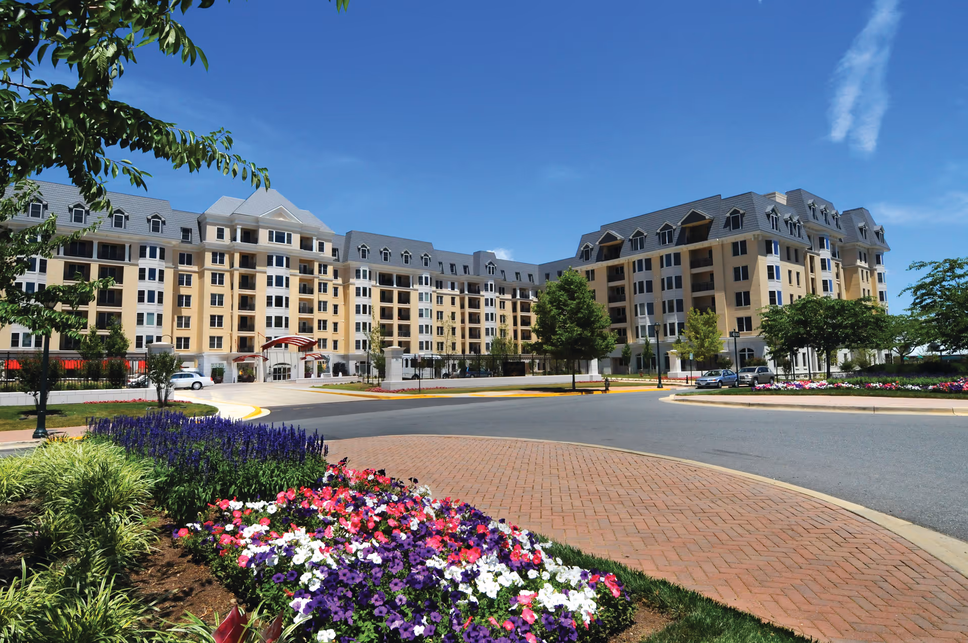 Exterior view of a large multi-story senior living facility building with beige and gray tones, surrounded by landscaped gardens with colorful flowers and green trees under a clear blue sky.