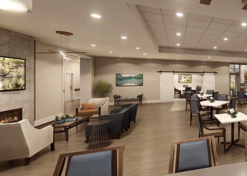 A spacious and well-lit common area in a senior living facility featuring a seating area with armchairs and a coffee table near a modern fireplace and a wall-mounted TV. The room has wooden flooring, neutral-colored walls, and ceiling lights. In the background, there are dining tables and chairs arranged neatly, separated by sliding barn doors. Decorative plants and landscape paintings adorn the walls.