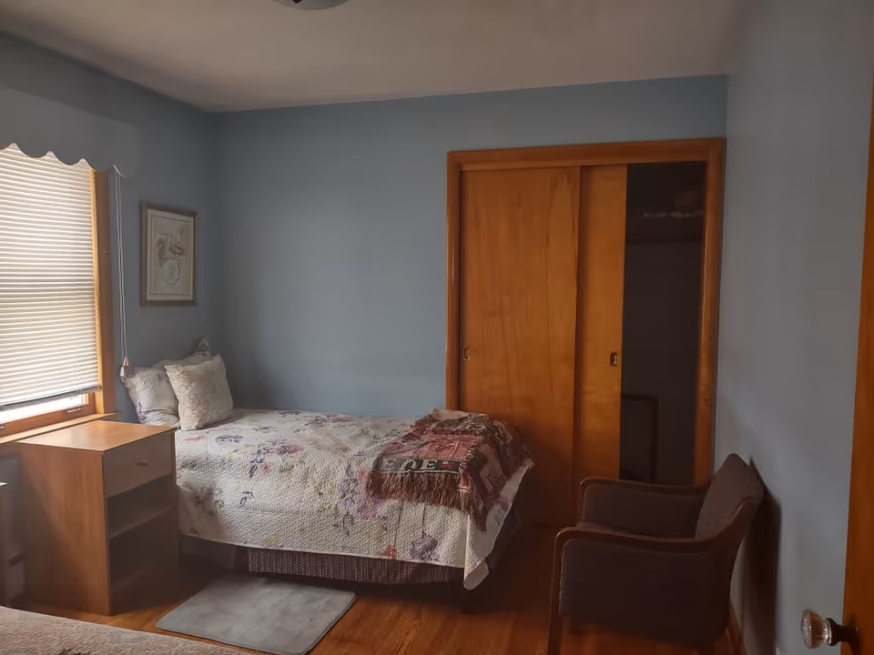A small bedroom with light blue walls, a window with closed blinds and a scalloped valance, a wooden nightstand, a bed with floral bedding and pillows, a folded blanket at the foot of the bed, a wooden sliding closet door, and a dark upholstered armchair.