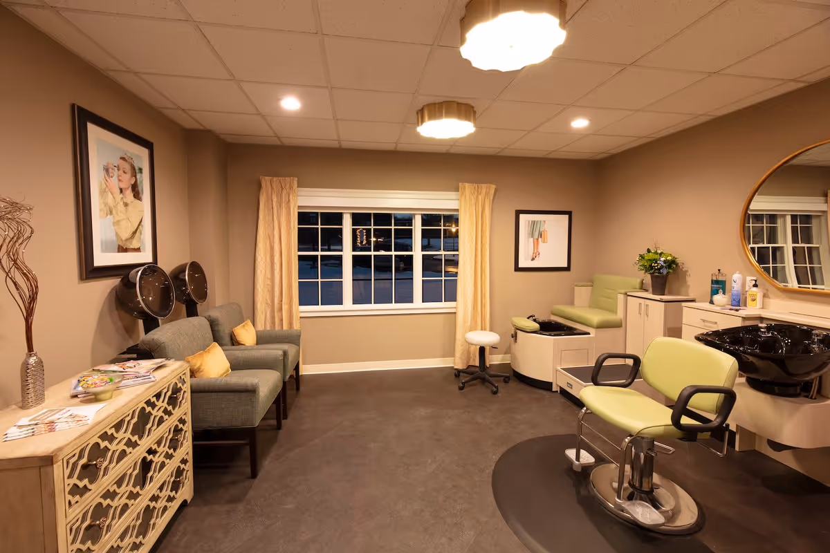 Well-lit salon room with a styling chair, shampoo sinks, hooded hair dryers, and a seating area by a window.