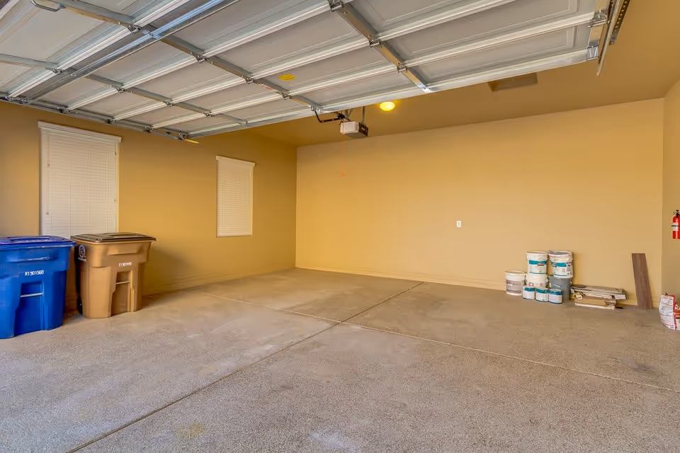 Empty two-car garage with the overhead door open, recycling and trash bins on the left and paint cans stacked on the right.