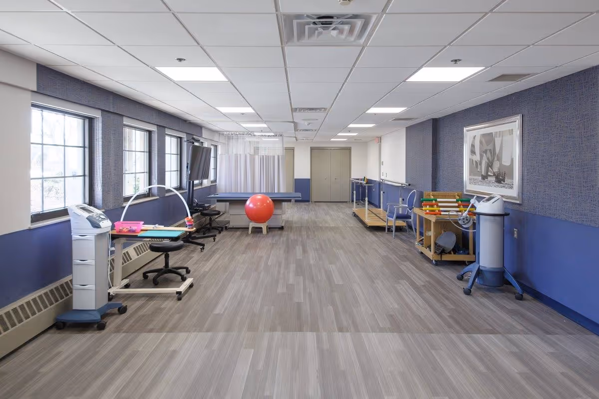 Large bright therapy room with exercise equipment, parallel bars, a red therapy ball, and windows along the left wall.