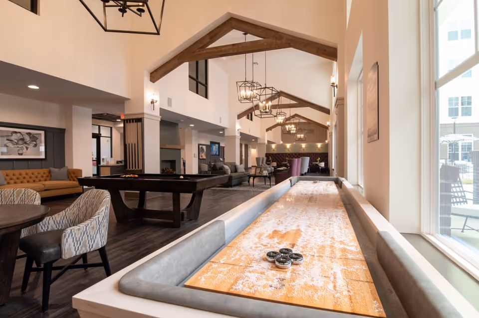 A spacious and well-lit common area in a senior living facility featuring a shuffleboard table in the foreground, a pool table in the middle, and various seating arrangements including sofas and chairs. The room has high ceilings with exposed wooden beams and modern hanging light fixtures. Large windows on the right side allow natural light to fill the space, and there is a view of an outdoor patio area through the windows.