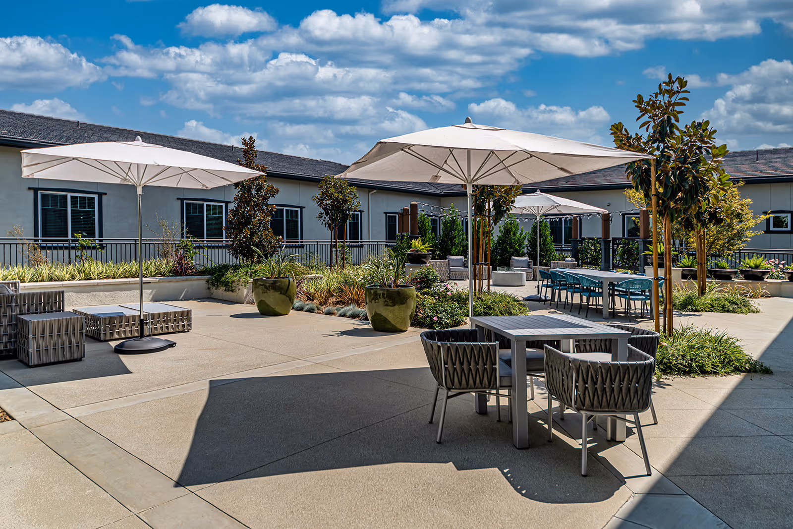 Outdoor patio area at Allara Senior Living with tables, chairs, large white umbrellas, potted plants, and surrounding building under a partly cloudy sky.