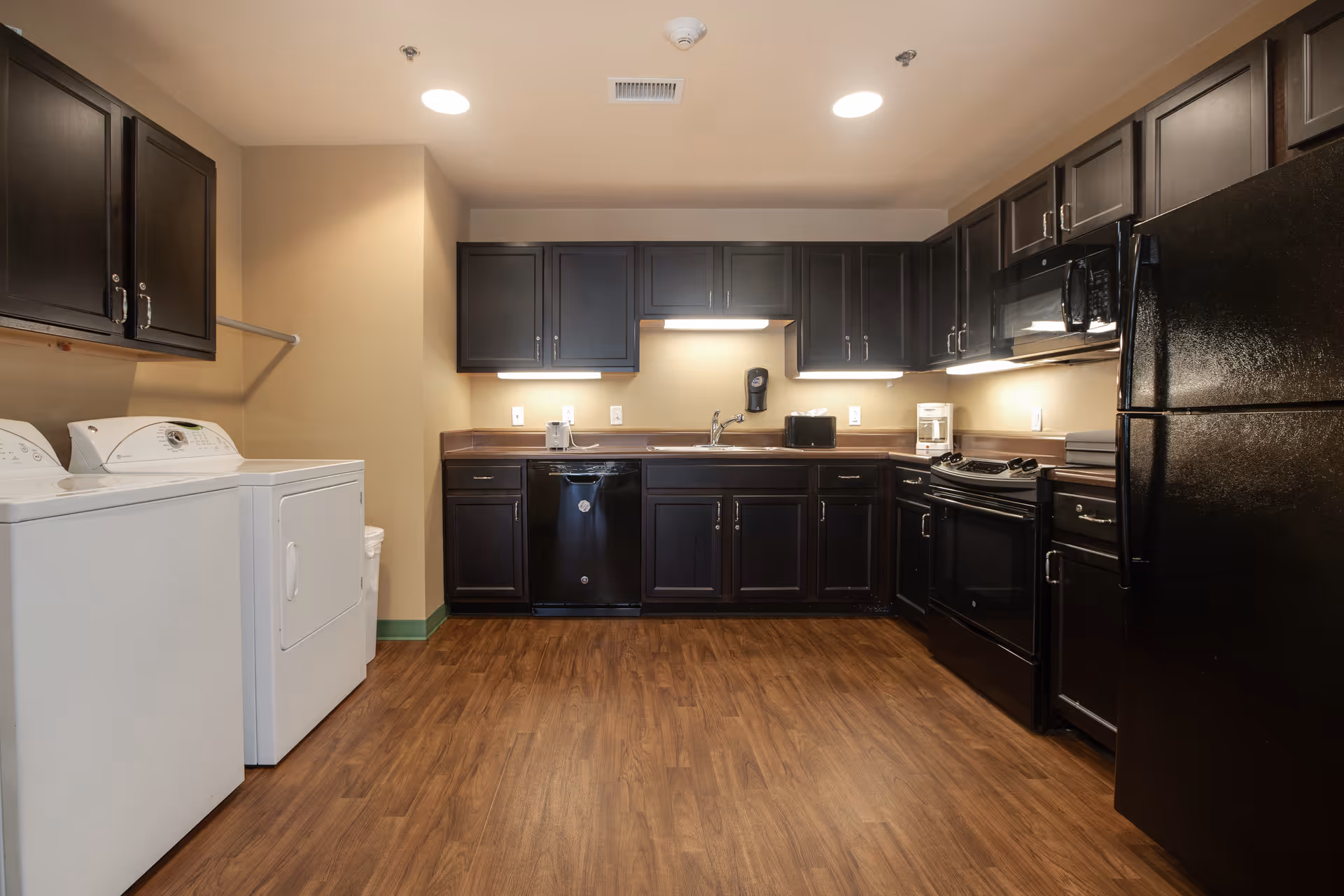 A kitchen area with dark wooden cabinets, black appliances including a refrigerator, stove, microwave, and dishwasher. There is a white washing machine and dryer on the left side. The floor is wooden and the walls are beige with under-cabinet lighting illuminating the countertop.