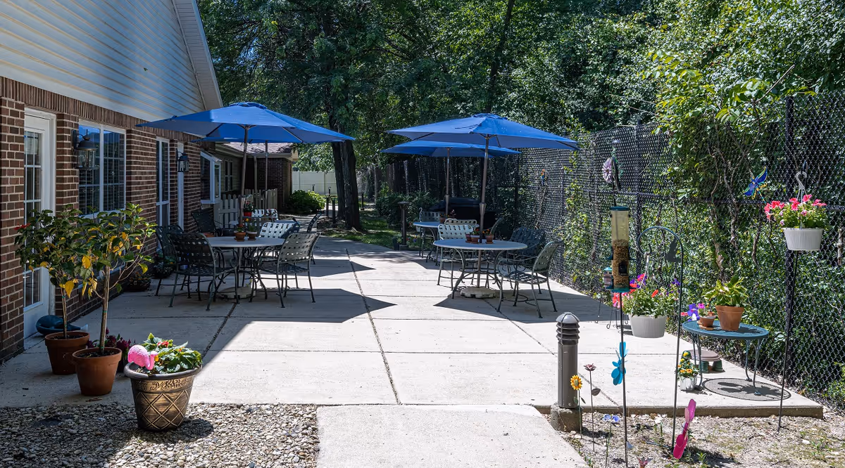 Outdoor patio area with round tables, chairs and blue umbrellas beside a brick building and chain-link fence, decorated with potted plants.