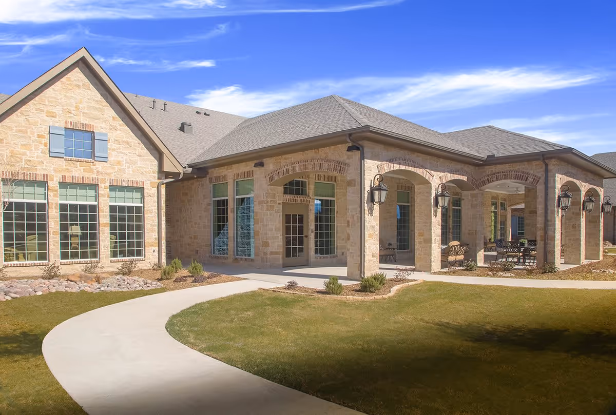 Exterior view of a senior living facility building with stone walls, large windows, and a covered patio area with outdoor seating. The building is surrounded by a well-maintained lawn and a curved concrete walkway under a blue sky with some clouds.