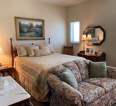 A cozy bedroom featuring a large bed with decorative pillows, a floral-patterned couch, a bedside table with a lamp, and a painting on the wall. Natural light enters through a window, creating a warm atmosphere.