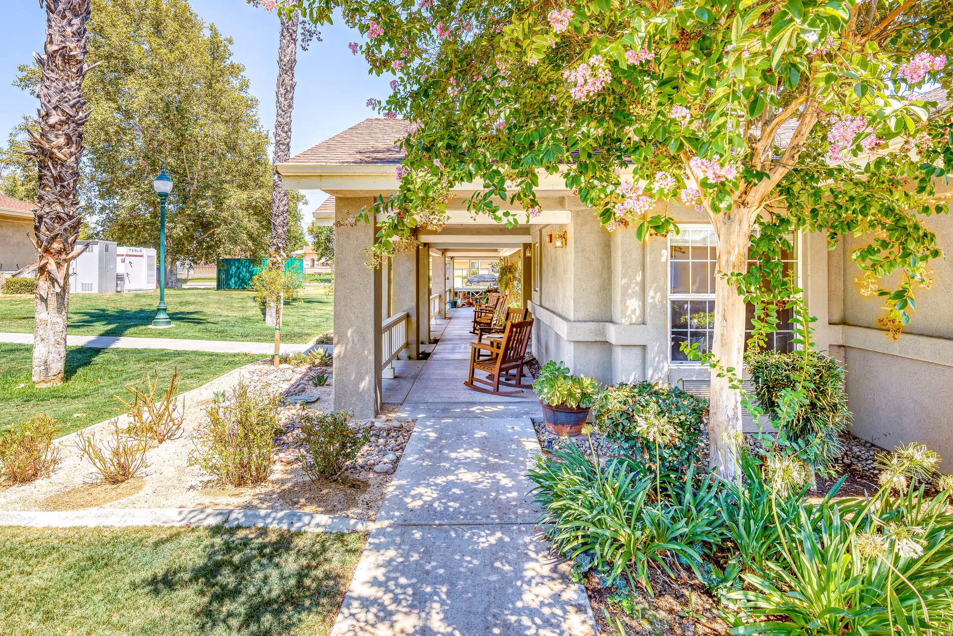 Covered exterior walkway with rocking chairs and landscaped greenery in front of a cottage building.
