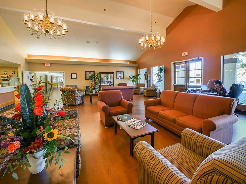 A spacious and well-lit living room area in a senior living facility with multiple comfortable chairs and sofas arranged around coffee tables. There are two chandeliers hanging from the ceiling, large windows letting in natural light, and a person sitting at a desk near the window working on a laptop. The room features warm colors with a mix of patterned and solid upholstery and decorative plants and flowers.