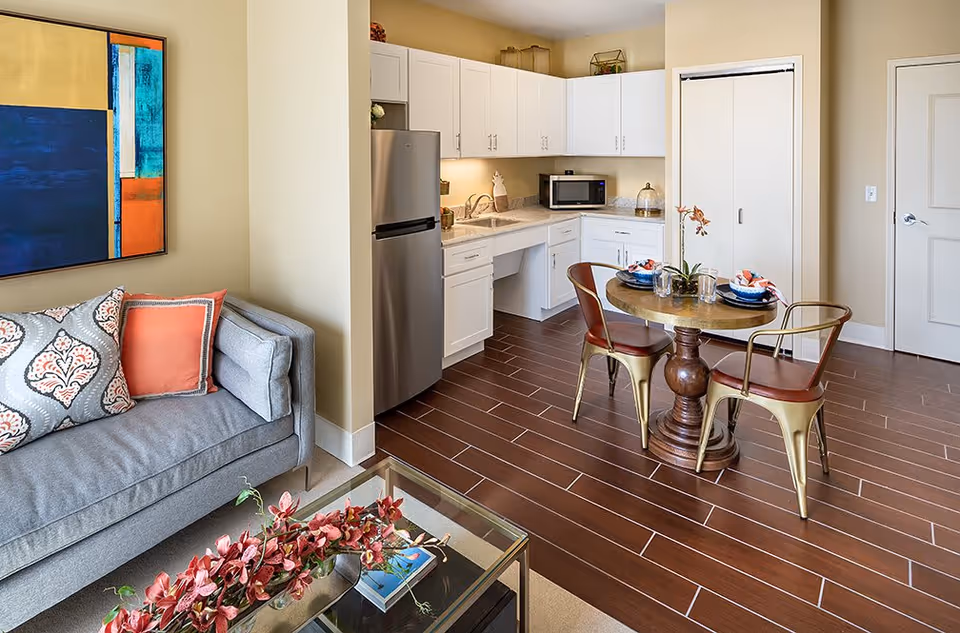 A bright and modern living space featuring a gray couch with decorative pillows, a glass coffee table with a floral arrangement, and a small round dining table set with two chairs. The kitchen area has white cabinets, a stainless steel refrigerator, a microwave, and a sink. The room has wood-look tile flooring and neutral-colored walls.