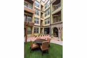 Outdoor courtyard area in a multi-story senior living facility with artificial grass, a round table, and six chairs. The building has multiple balconies and windows overlooking the courtyard.
