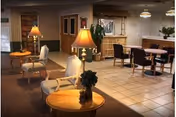 Cozy senior living facility common area with armchairs, table lamps, round dining tables and a reception desk.
