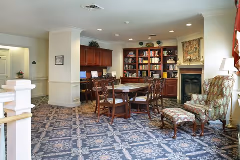 Communal lounge with a central table and chairs, bookshelves, a fireplace, and an upholstered armchair.
