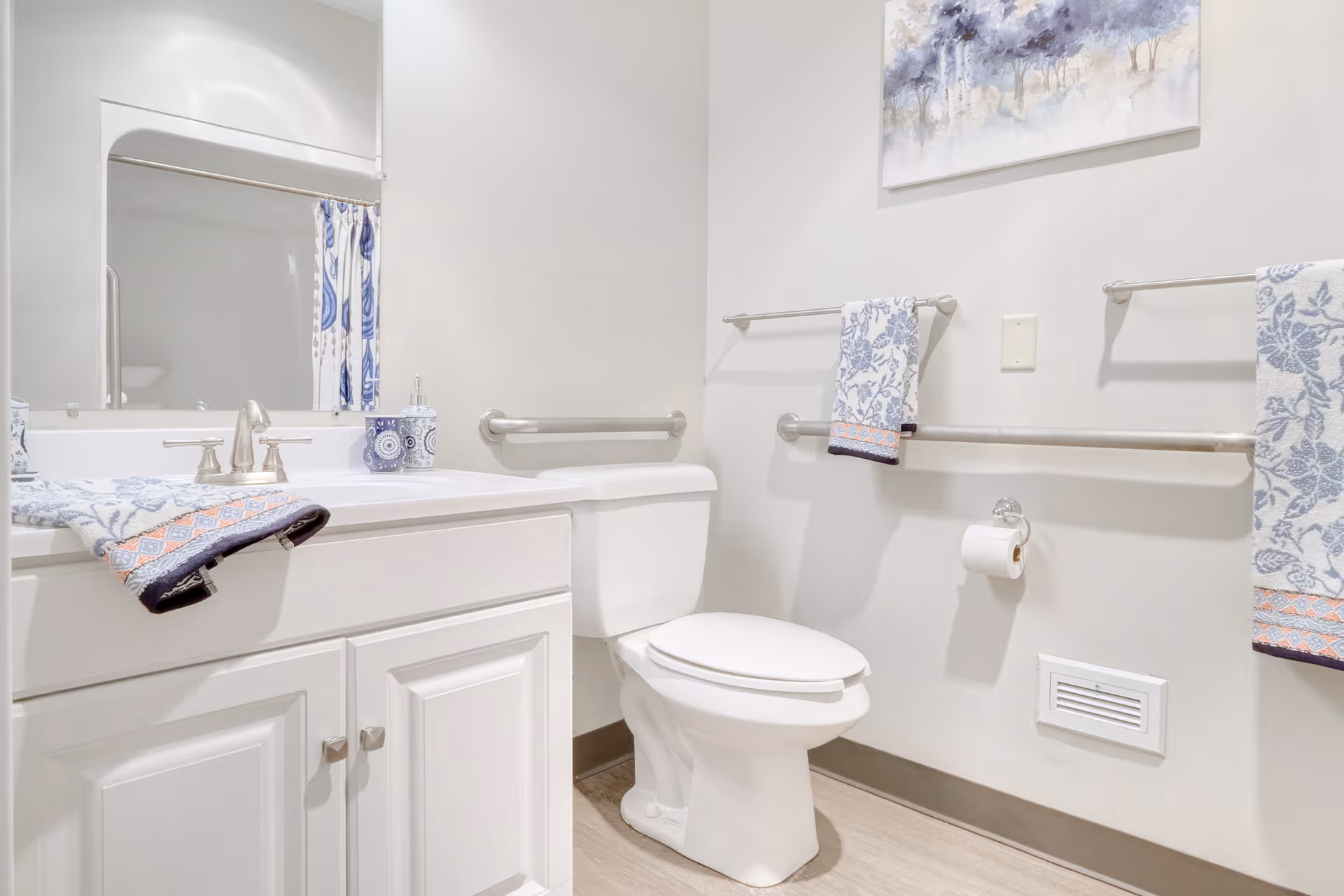 A clean and bright bathroom featuring a white toilet, a white vanity with a sink and silver faucet, a large mirror above the sink, and multiple grab bars on the walls. There are decorative blue and white towels hanging on the grab bars and a matching soap dispenser on the vanity. A piece of artwork with a nature theme hangs on the wall above the toilet.