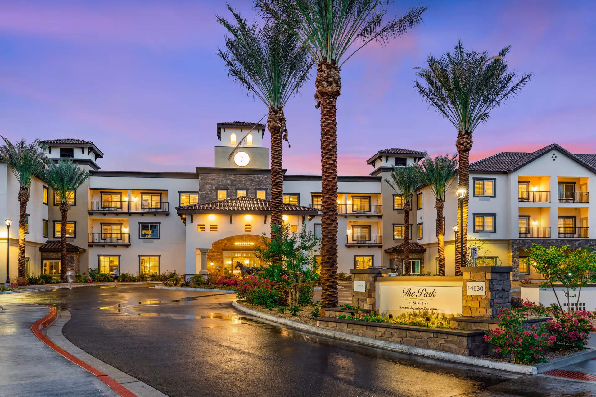 Exterior view of The Park at Surprise Independent Living Community building at dusk, featuring a well-lit entrance, palm trees, and a sign with the community name and address 14630.