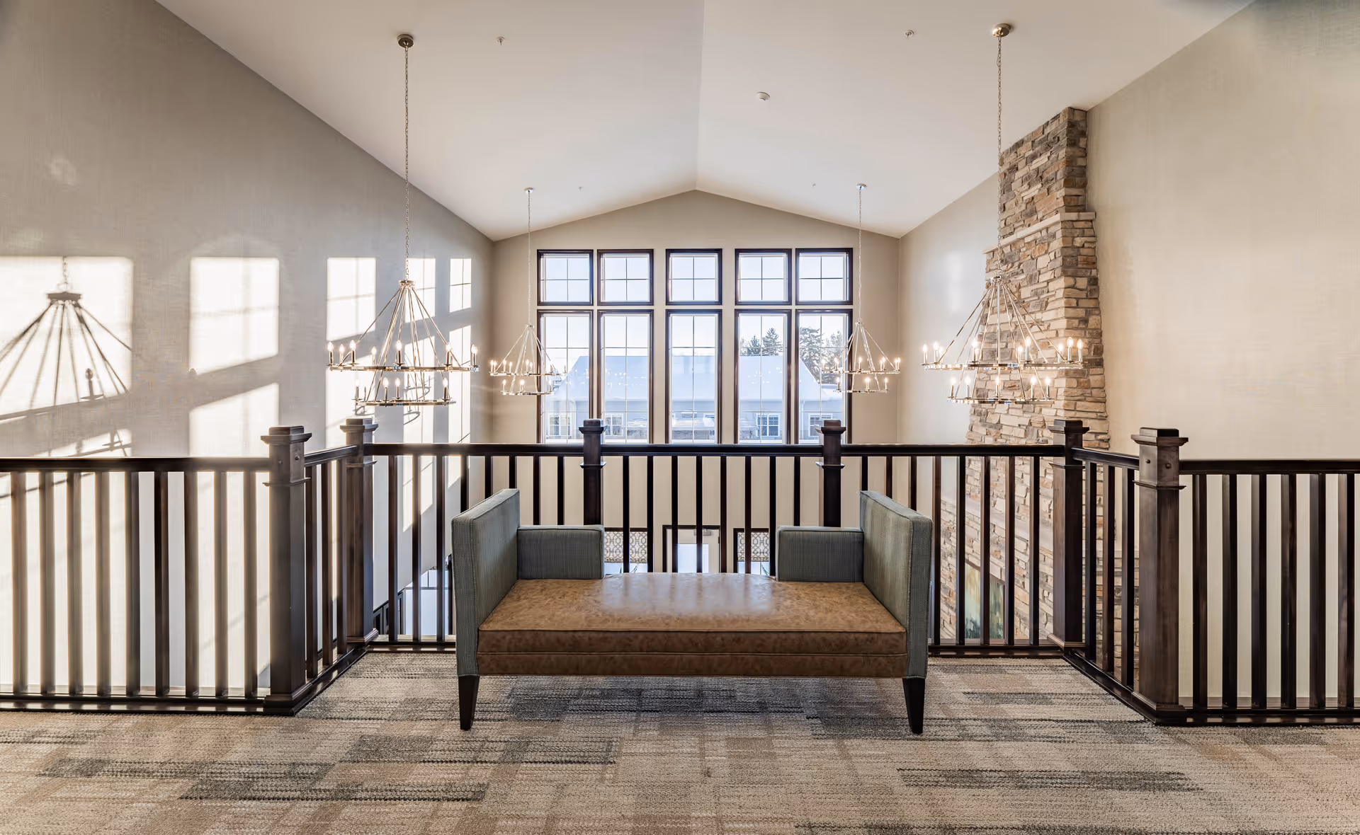 A spacious interior area with a high vaulted ceiling, large windows letting in natural light, and a stone fireplace on the right. A modern bench with green upholstered sides and a brown seat is positioned in the center, surrounded by dark wooden railings. Elegant chandeliers hang from the ceiling, casting shadows on the walls.