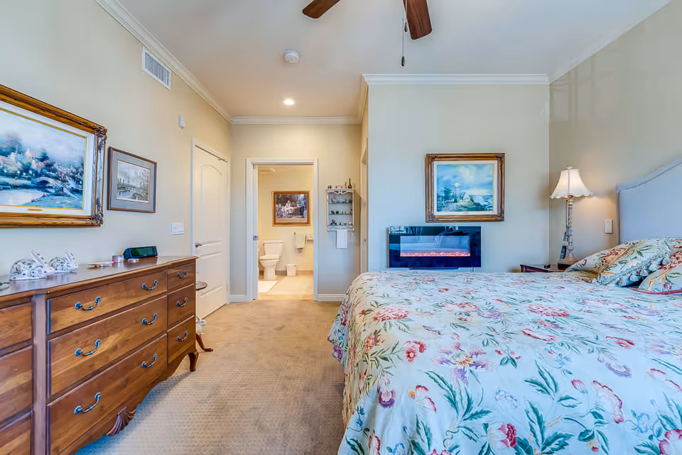 A cozy bedroom with a floral bedspread on a large bed, a wooden dresser with decorative items, framed paintings on the walls, a ceiling fan, and a small electric fireplace mounted on the wall. The room has beige walls and carpet, and an open door reveals a bathroom with a toilet and framed artwork.