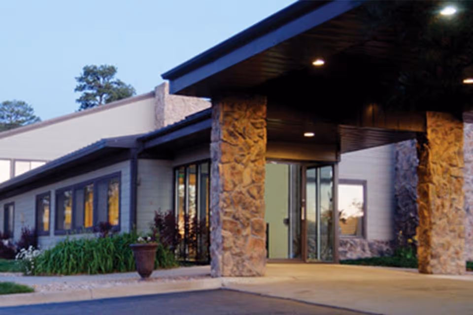 Entrance of a building with stone pillars supporting a covered driveway, large windows along the side, and a glass door entrance. There are plants and greenery near the building and a clear sky above.