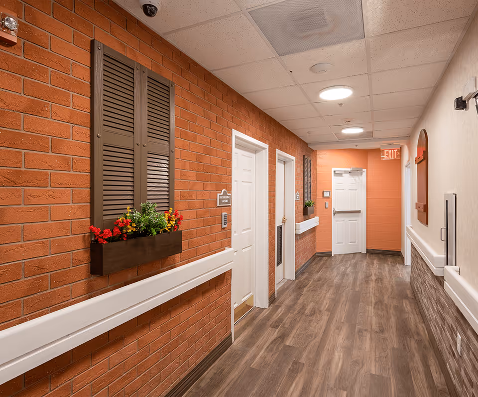 A well-lit hallway in a senior living facility with brick and beige walls, wood-look flooring, white doors, and decorative window shutters with flower boxes. There are handrails along the walls and an exit sign at the end of the corridor.