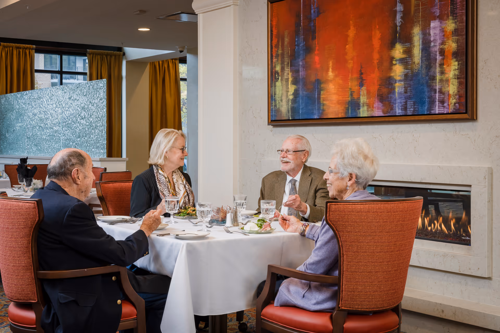 Four older adults seated around a white-clothed dining table in an elegant dining room by a fireplace and colorful abstract painting.