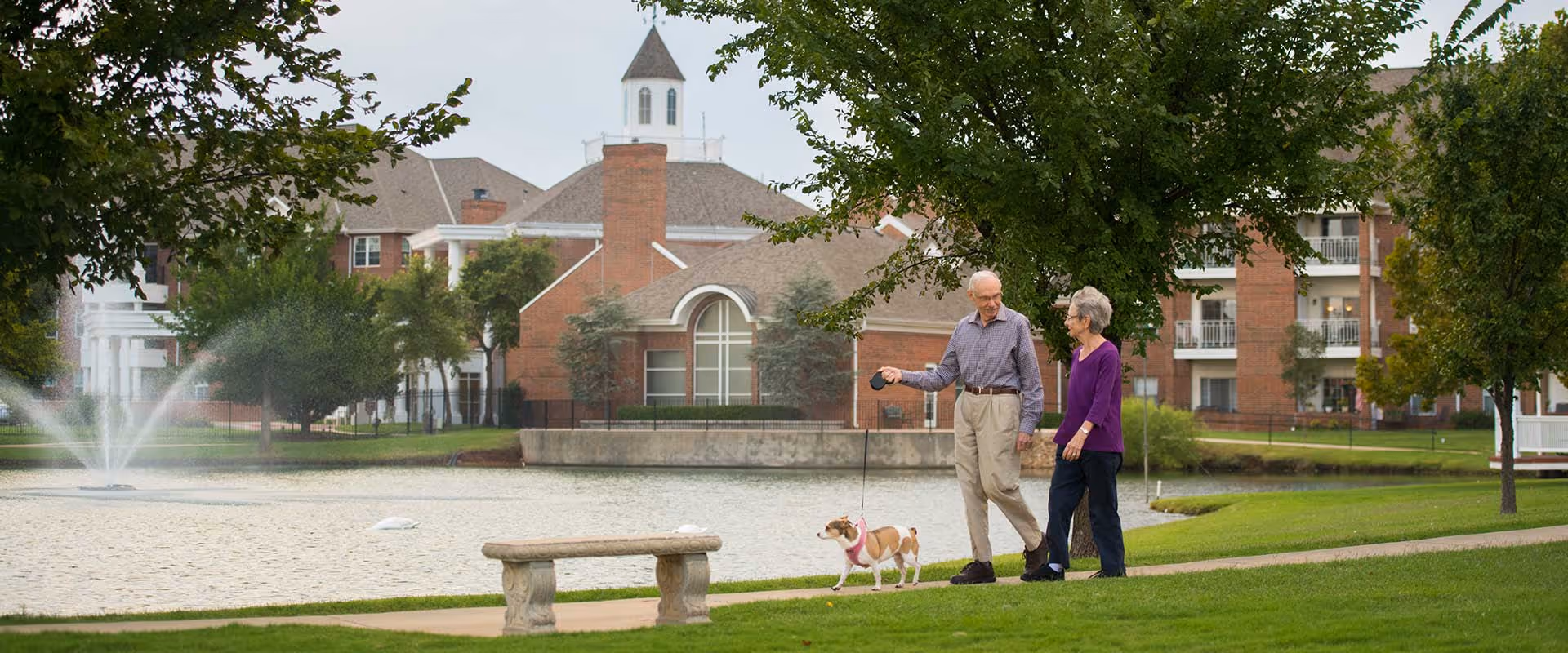 An elderly couple walking a small dog on a leash along a paved path beside a pond with a fountain, surrounded by green grass and trees, with a large brick building in the background.