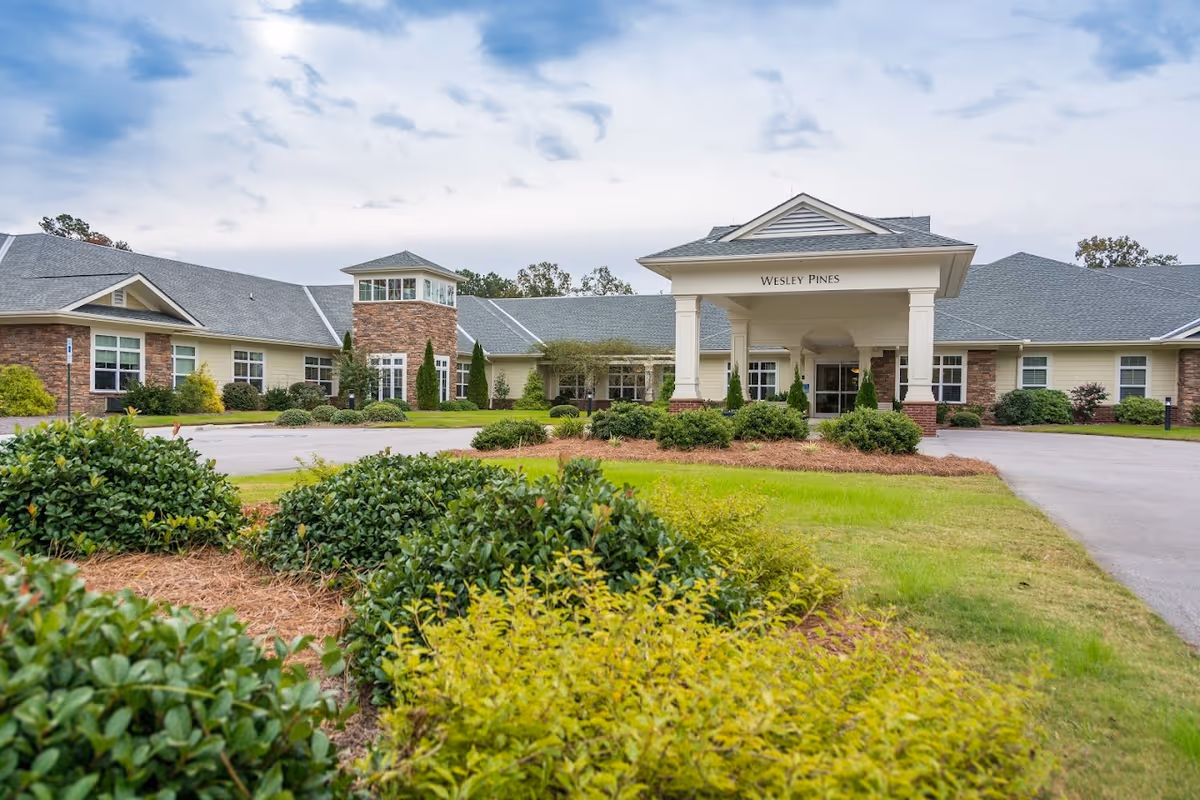 Exterior view of Wesley Pines Retirement Community building with a covered entrance, surrounded by well-maintained landscaping including bushes and grass under a partly cloudy sky.