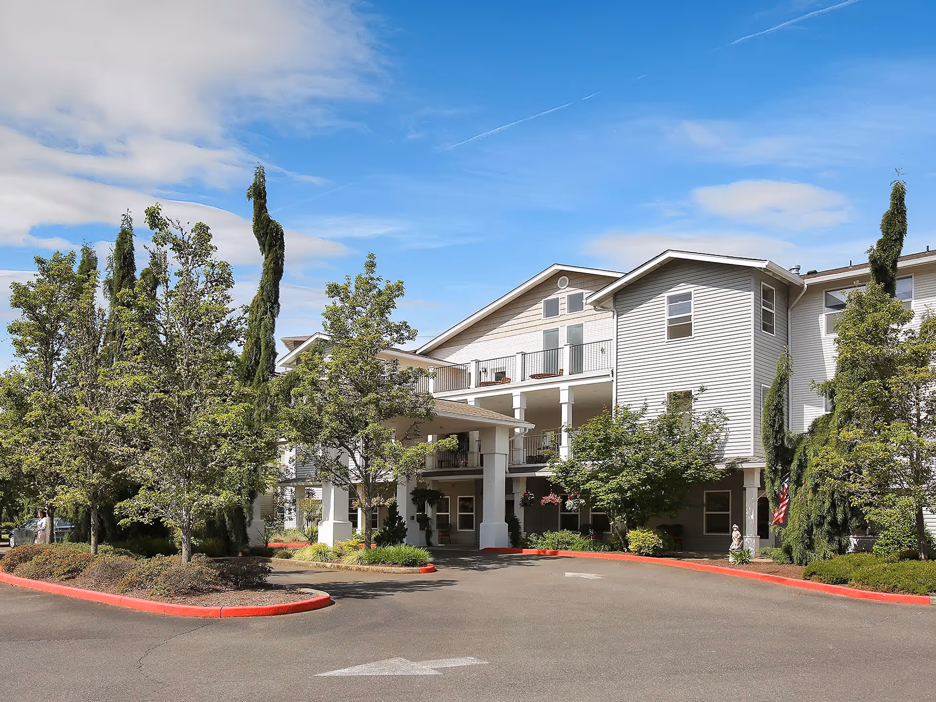 Exterior view of a senior living facility building with a covered entrance, surrounded by trees and landscaping under a partly cloudy blue sky.