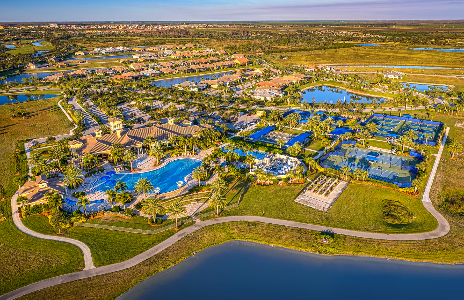 Aerial view of Del Webb Naples 55+ Retirement Community showing a large swimming pool surrounded by palm trees, multiple tennis and pickleball courts, walking paths, residential homes, and landscaped green areas with water features.
