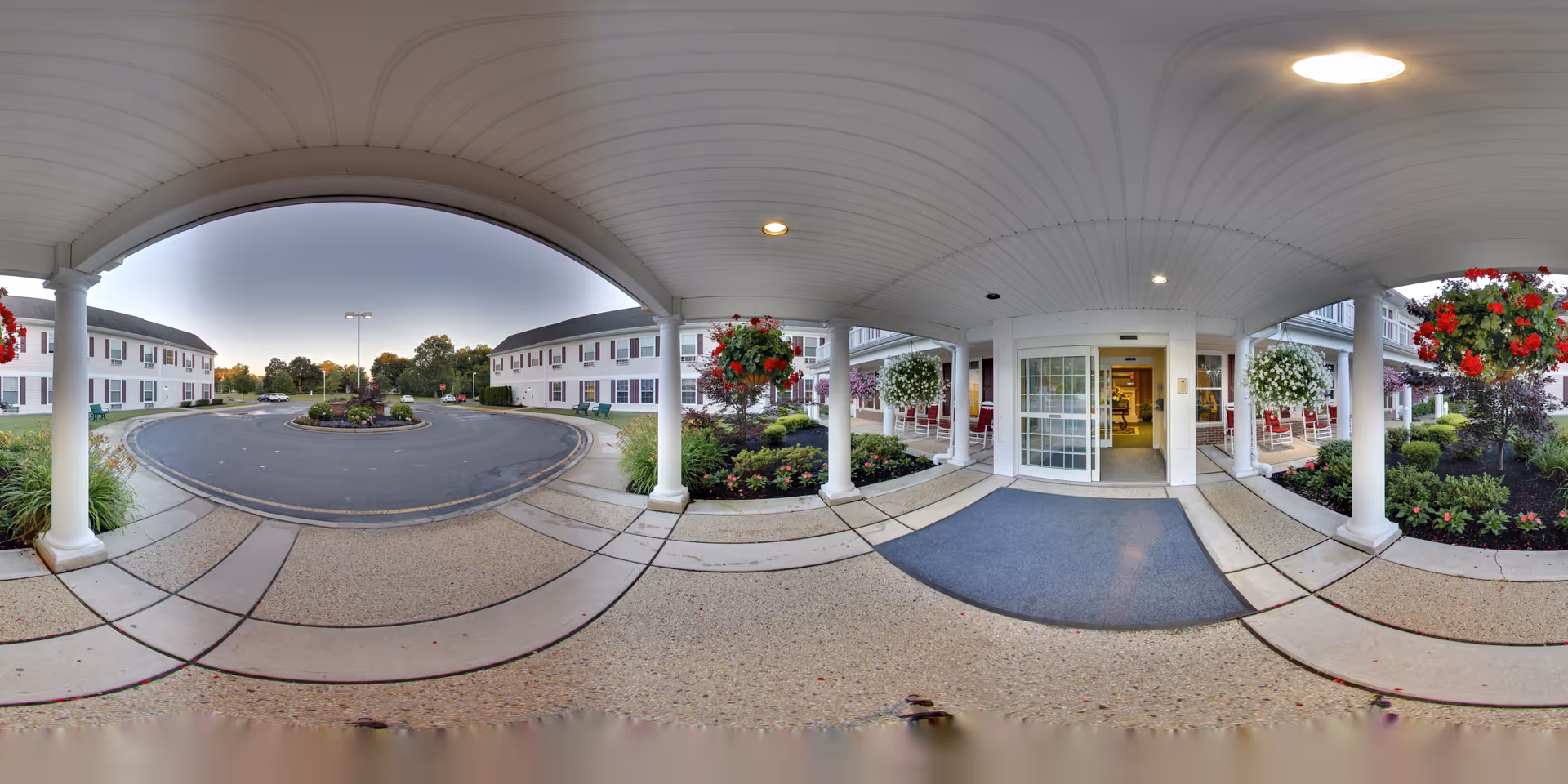 Covered entrance area of Brandywine Toms River by Monarch with white pillars, hanging flower baskets, and a view of the driveway and surrounding buildings under a clear sky at dusk.